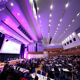 Der Maria-Theresia-Saal aus einem anderen Blickwinkel. Viele Gäste sitzen in dem Saal, der in leuchtenden Farben angestrahlt wird. Der Blick geht zur Bühne links. Foto: Alexander Kaya